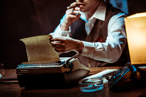 Cropped view of detective in glasses smoking cigar while using typewriter in dark office