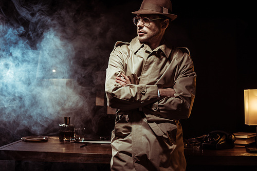 Detective in trench coat and hat standing with crossed arms in office