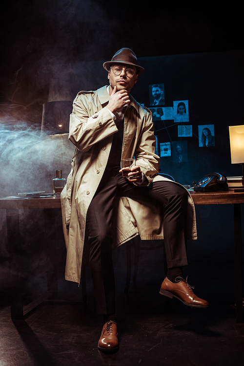 Detective in trench coat and hat sitting on table and holding glass of cognac