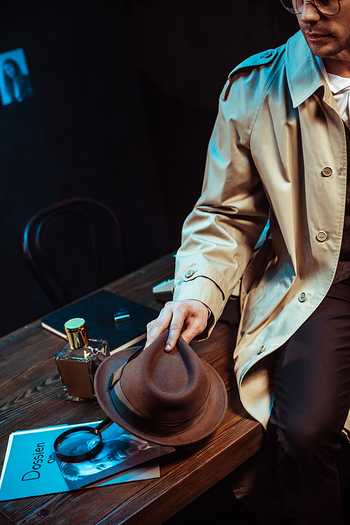 Cropped view of detective in trench coat sitting on table and holding hat