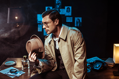 Pensive detective in glasses and trench coat sitting on table and holding hat