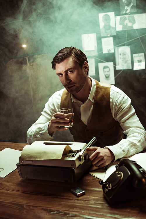 Detective holding glass of cognac while using typewriter in dark office