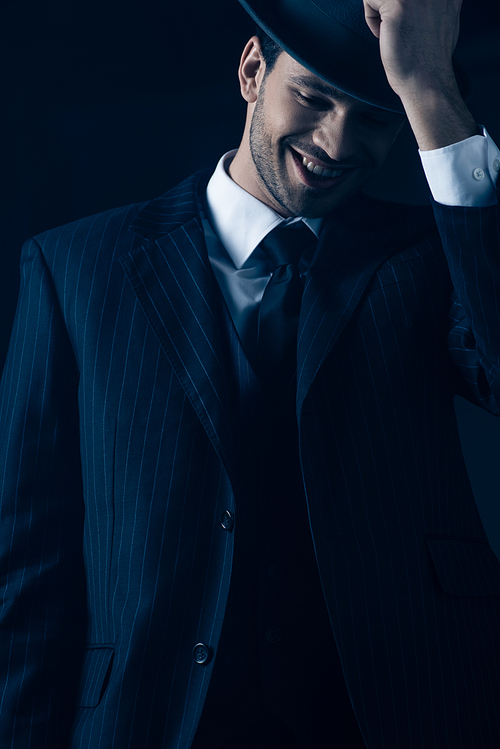 Front view of mafioso holding felt hat and laughing on dark background