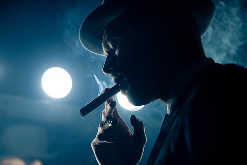 Low angle view of mafioso smoking cigar on dark blue background