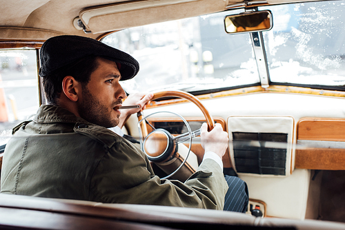 Selective focus of mafioso in coat and hat with cigarette holding steering wheel in car