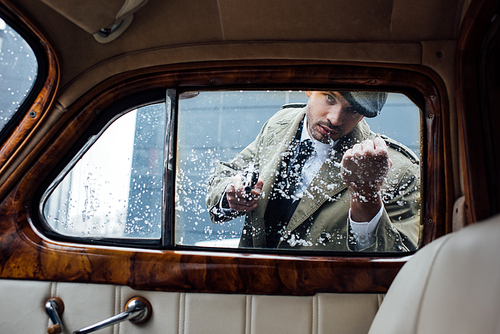 Angry mafioso aiming gun, showing fist and looking into retro car