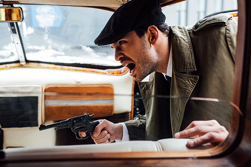 Selective focus of angry mafioso aiming gun and shouting in retro car