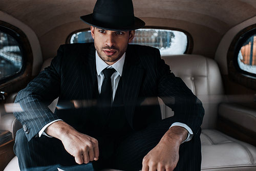 Selective focus of pensive mafioso in black suit and felt hat in retro car