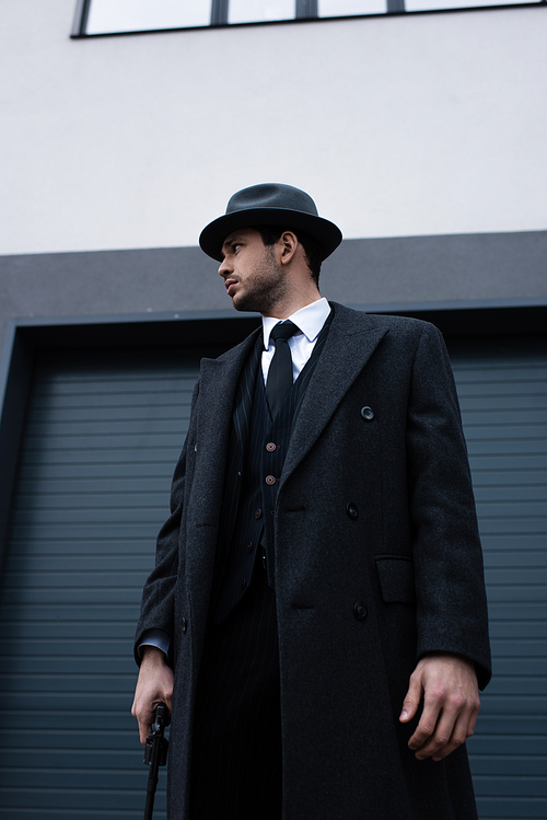 Low angle view of gangster in black coat and hat with revolver looking away on street