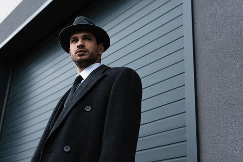 Low angle view of mafioso in black coat and felt hat near wall on street