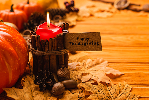 selective focus of burning candle with happy thanksgiving card and autumnal decoration on wooden background