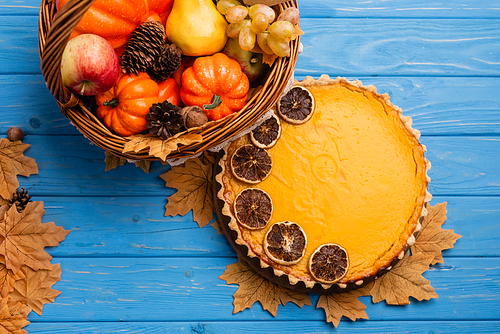 top view of autumnal harvest in wicker basket with happy thanksgiving card near pumpkin pie on blue wooden background