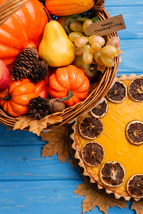 top view of autumnal harvest in wicker basket with happy thanksgiving card near pumpkin pie on blue wooden background