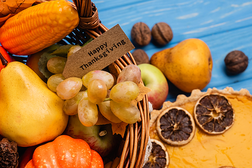 close up view of autumnal harvest in wicker basket with happy thanksgiving card near pumpkin pie on blue wooden background