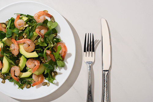 top view of fresh green salad with pumpkin seeds, shrimps and avocado on plate near cutlery on white background