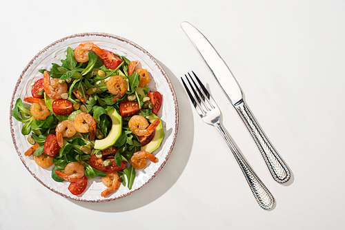 top view of fresh green salad with shrimps and avocado on plate near cutlery on white background