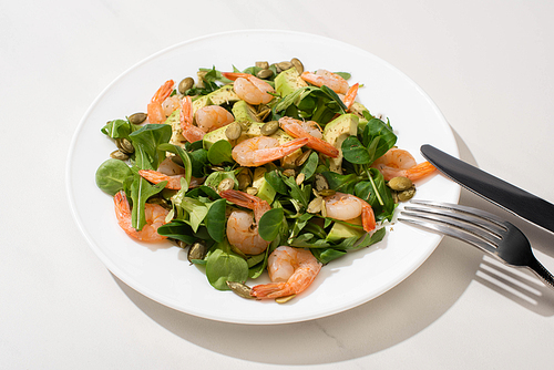 fresh green salad with pumpkin seeds, shrimps and avocado on plate near cutlery on white background