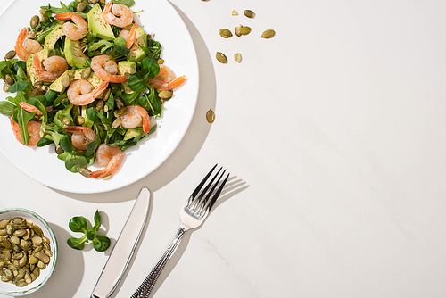 top view of fresh green salad with pumpkin seeds, shrimps and avocado on plate near cutlery on white background