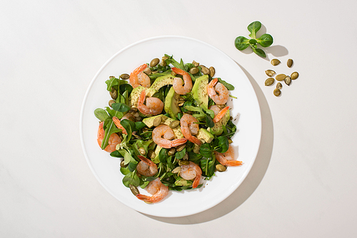top view of fresh green salad with pumpkin seeds, shrimps and avocado on plate on white background
