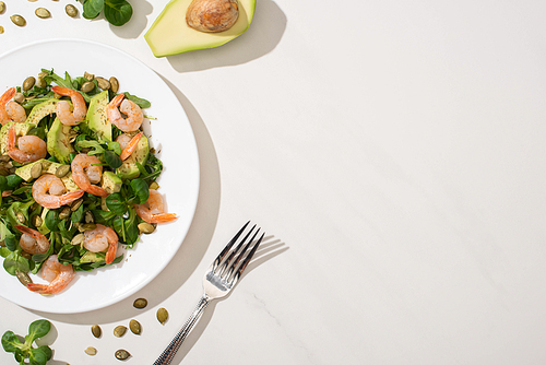 top view of fresh green salad with pumpkin seeds, shrimps and avocado on plate near fork and ingredients on white background