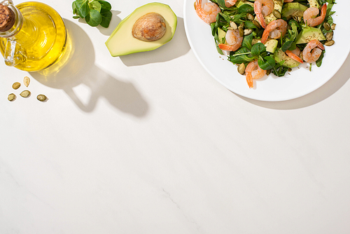 top view of fresh green salad with pumpkin seeds, shrimps and avocado on plate near ingredients on white background