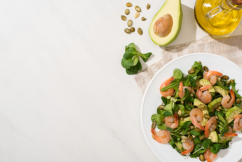 top view of fresh green salad with pumpkin seeds, shrimps and avocado on plate on napkin near ingredients on white background