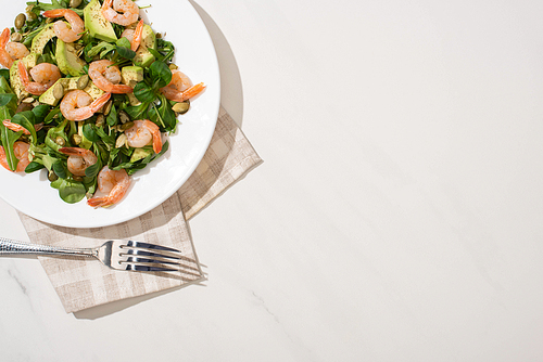 top view of fresh green salad with pumpkin seeds, shrimps and avocado on plate on napkin near fork on white background