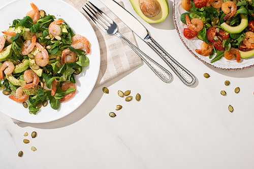 top view of fresh green salad with shrimps and avocado on plates near cutlery on white background