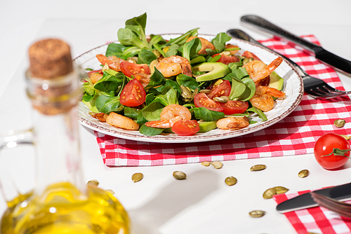 selective focus of fresh green salad with shrimps and avocado on plate near cutlery on plaid napkin and jar of oil on white background