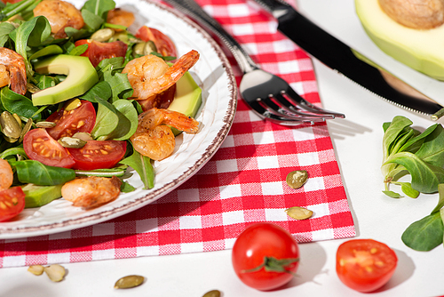 close up view of fresh green salad with shrimps and avocado on plate near cutlery on plaid napkin and ingredients on white background