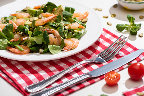 fresh green salad with shrimps and avocado on plate near cutlery on plaid napkin and ingredients on white background
