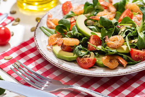 selective focus of fresh green salad with shrimps and avocado on plate near cutlery on plaid napkin