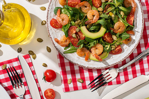 top view of fresh green salad with shrimps and avocado on plate near cutlery on plaid napkin and ingredients on white background