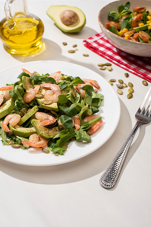 selective focus of fresh green salad with shrimps and avocado on plates near fork and oil