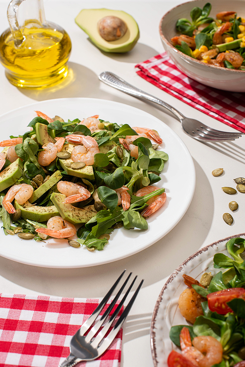 selective focus of fresh green salad with shrimps and avocado on plates near forks and oil