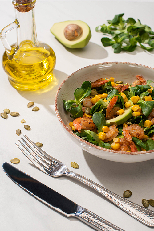 fresh green salad with corn, shrimps and avocado on plate near cutlery on white background