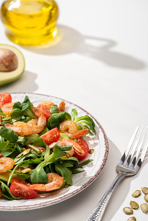 selective focus of fresh green salad with shrimps and avocado on plate near fork and ingredients on white background
