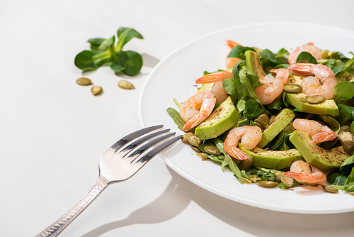 selective focus of fresh green salad with shrimps and avocado on plate near fork on white background