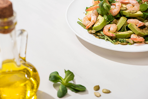 selective focus of fresh green salad with shrimps and avocado on plate near olive oil on white background