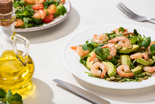 selective focus of fresh green salad with shrimps and avocado on plate near olive oil and cutlery on white background