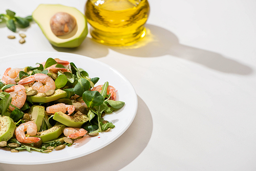 selective focus of fresh green salad with shrimps and avocado on plate near olive oil on white background