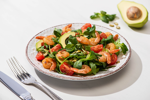 selective focus of fresh green salad with shrimps and avocado on plate near cutlery on white background