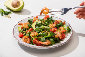 partial view of woman eating fresh green salad with cherry tomatoes, shrimps and avocado on white background