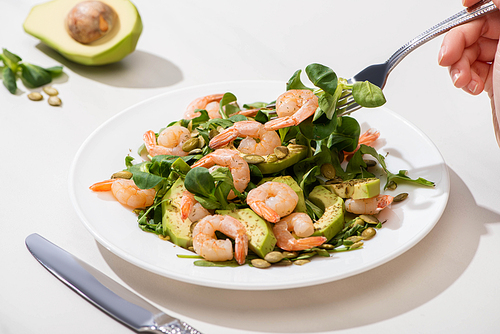 partial view of woman eating fresh green salad with pumpkin seeds, shrimps and avocado on white background