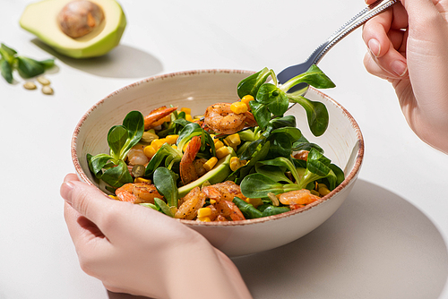 partial view of woman eating fresh green salad with corn, shrimps and avocado on white background
