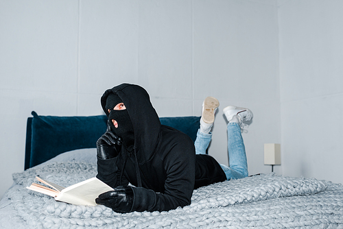 Pensive robber in balaclava holding book while lying on bed