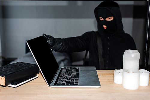 Selective focus of thief in balaclava taking laptop from table