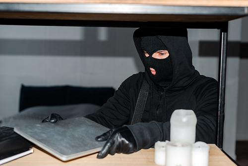 Selective focus of robber in balaclava taking laptop from cupboard