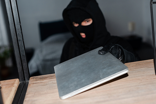 Selective focus of robber in balaclava and leather glove taking laptop from cupboard