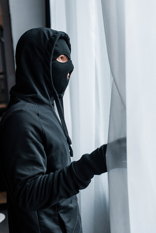 Side view of robber in balaclava standing near curtains